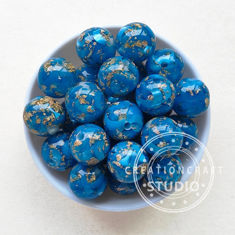 May include: A bowl of teal blue and gold beads. The beads are round and have a marbled effect with gold flecks throughout.
