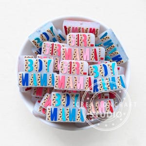 May include: A bowl filled with colourful resin charms with the words "Boy", "Girl", and "Mama" in various colours and patterns. The charms are decorated with glitter and a leopard print design.