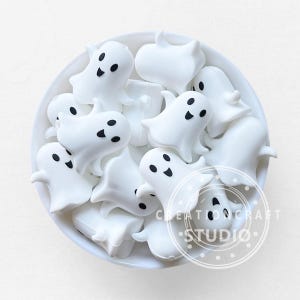 May include: A white bowl filled with small, white ghost-shaped decorations. Each ghost has black dot eyes and a smiling mouth. The bowl has the text "CREATION CRAFT STUDIO" in a circular design.