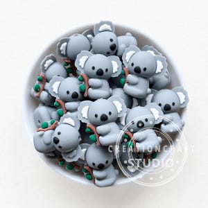 Puede incluir: Un bol lleno de cuentas de silicona grises con forma de koala. Las cuentas tienen pequeñas ramas marrones y hojas verdes unidas a ellas. Las cuentas están diseñadas para el juego sensorial y la dentition.
