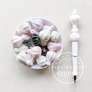 Puede incluir: Un tazón blanco lleno de cuentas de vidrio iridiscentes blancas y rosadas con forma de bulbo de ajo. Un bolígrafo blanco con un diseño de cuentas rosas y blancas está al lado del tazón. El texto "CREATIONCRAFT STUDIO" es visible en la parte inferior derecha de la imagen.