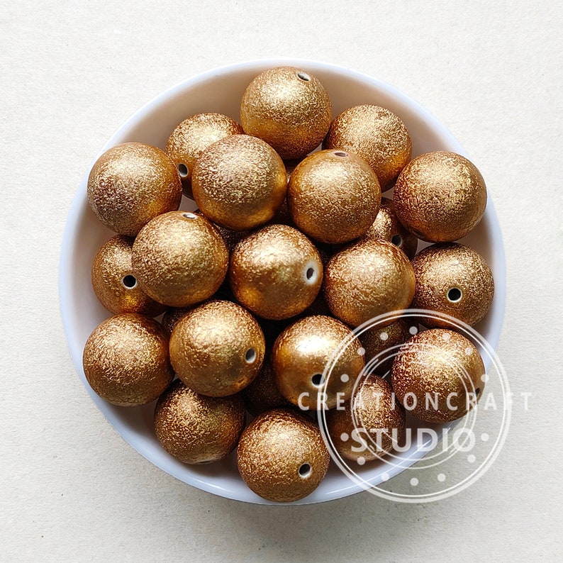 May include: A white bowl filled with gold glitter beads. The beads are round and have a textured surface.