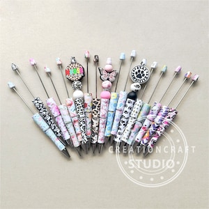 May include: A collection of 18 colorful pens with various designs and patterns. The pens feature different toppers, including a butterfly, a paw print, and a "Nurse" charm. The pens are arranged in a fan shape on a light brown surface.