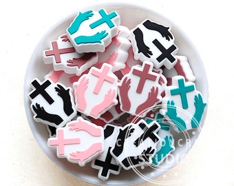 May include: A bowl filled with colorful silicone charms featuring a cross and hands in a praying position. The charms are in various colors, including pink, teal, black, and brown.