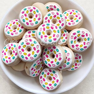 Op de afbeelding: Een kom gevuld met donutvormige siliconen kralen. De kralen zijn wit met roze, gele en turquoise sprinkles.