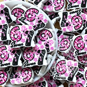May include: A collection of white, pink, and black decorative embellishments. Each piece features the number "67" with a pink leopard print design, a pink bow, and the word "Seven". The embellishments have a zebra-striped pattern and star accents.