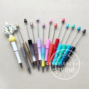 May include: A collection of 13 colourful pens with different coloured tops and metallic accents. The pens have a variety of designs, including a bee with the text "Be Your Wild, Be Humble, Be True".
