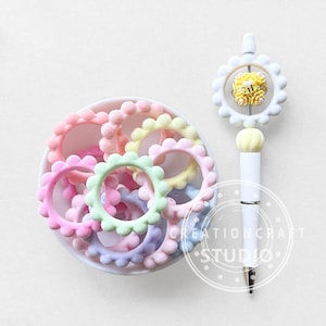 May include: A collection of colorful hair ties in a white bowl, alongside a white pen with a flower-shaped top. The hair ties are in shades of pink, green, yellow, and purple. The pen has a yellow flower design and the text "CREATIONCRAFT STUDIO".