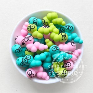 Puede incluir: Un bol lleno de cuentas de gusano de silicona de colores. Los gusanos son verdes, rosas y azules y tienen ojos negros y una boca sonriente. Las cuentas son perfectas para hacer collares, pulseras u otras manualidades.