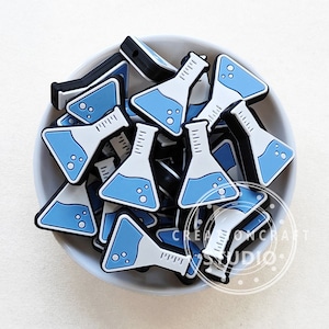 Pode incluir: Uma tigela cheia de pingentes de béquer de silicone azul e branco. Os béqueres têm uma base branca e um líquido azul com bolhas dentro. Os pingentes são perfeitos para adicionar um tema científico a joias ou artesanato.