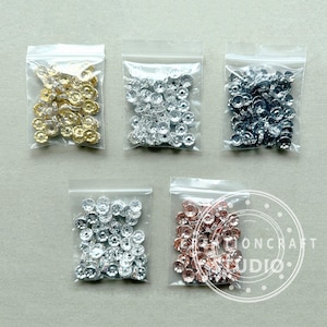 May include: Five small clear plastic bags each containing a different color of rhinestone embellishments. The colors include gold, silver, dark gray, clear, and rose gold. The rhinestones are round with a flat back and a faceted front.