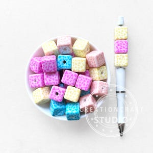 May include: A selection of colourful square beads in a white bowl, next to a white pen with matching beads. The beads are in shades of pink, yellow, blue, and peach, with a heart-shaped pattern. The pen has a silver accent.