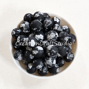 May include: A collection of black round beads with a matte finish, each adorned with a delicate white and gray floral pattern. The beads are piled in a white bowl, creating a visually appealing contrast. The text "CreationCraftStudio" is visible.