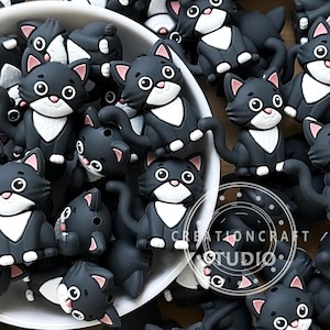 Peut inclure: Un ensemble d'ornements en forme de chat noir avec des plastrons blancs, des oreilles roses et de grands yeux ronds. Les figurines de chat sont regroupées, suggérant un matériel de bricolage ou un objet décoratif. Le texte "CREATIONCRAFT STUDIO" est inclus.