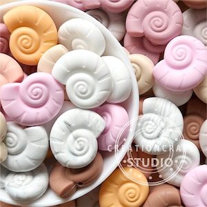 May include: A collection of colourful silicone seashell-shaped beads. The beads are in shades of pink, white, brown, and orange. The beads are perfect for making necklaces, bracelets, or other jewellery.