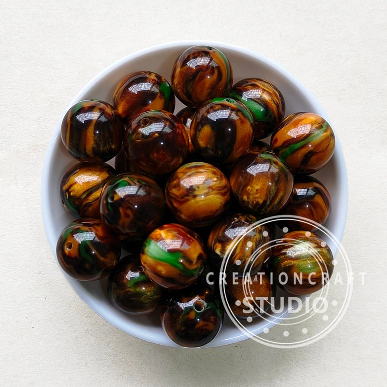 Op de afbeelding: Een kom met ronde, bruine, groene en gouden marmerachtige kralen. De kralen zijn ongeveer 2,5 cm in diameter en hebben een glanzende afwerking.
