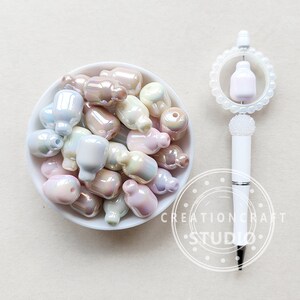 May include: A white bowl filled with iridescent pastel-colored glass beads shaped like bottles. The beads have a pearlescent finish and are arranged in a random pattern. The bowl is sitting on a white surface.