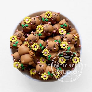 Puede incluir: Un conjunto de figuritas de wombat marrones, cada una sosteniendo una flor amarilla con hojas verdes. Las figuritas están en un cuenco blanco. El texto "CREATIONCRAFT STUDIO" es visible.