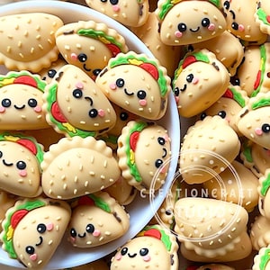 Pode incluir: Uma coleção de amuletos em forma de taco bege com rostos sorridentes, detalhes em alface verde e tomate vermelho. Os amuletos estão espalhados em uma tigela branca. O texto "CREATIONCRAFT STUDIOS" é visível.