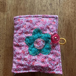Puede incluir: Una bolsa de tela rosa con un estampado floral y una aplicación de flor verde. La bolsa tiene un botón rojo y un lazo elástico amarillo.