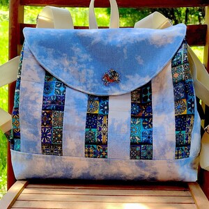 Könnte beinhalten: Ein blauer und weißer gestreifter Rucksack mit einer blauen und weißen gemusterten Klappe. Der Rucksack hat einen weißen Riemen und ein dekoratives Metallstück auf der Klappe.