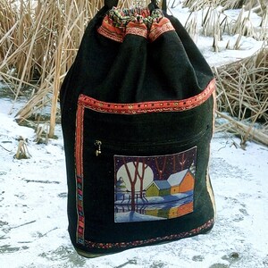 Pode incluir: Saco preto de cordão com uma cena de inverno bordada na frente. A cena apresenta uma lua branca, duas árvores, uma casa e um rio. O saco tem guarnições vermelhas, laranja e douradas.