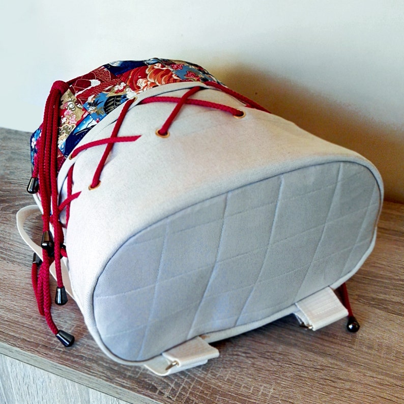 May include: A grey drawstring backpack with red laces and a floral pattern on the top flap. The backpack has a quilted bottom and white straps.