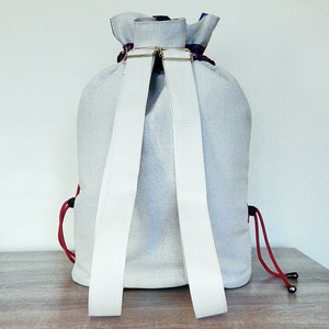 May include: A white canvas backpack with red drawstrings and white straps. The backpack has a gold metal buckle.