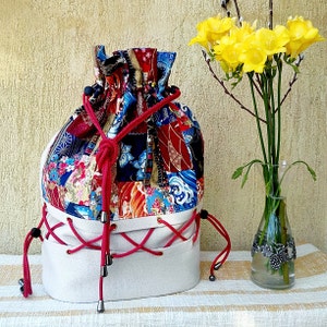 May include: A white drawstring bag with red laces and a colourful patchwork design. The bag is made of fabric and has a round bottom. The bag is sitting on a white surface with a bouquet of yellow flowers in a glass vase.