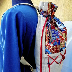 May include: A beige drawstring backpack with red and white drawstrings. The backpack is made of a canvas material and has a colourful patchwork design on the front. The design features a variety of geometric shapes, flowers, and other patterns in shades of red, blue, and gold.