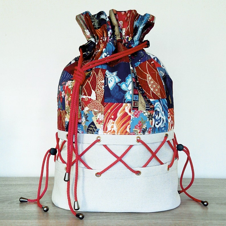 May include: A large, white drawstring bag with red cords. The bag is made of canvas and has a patchwork design of colourful fabrics with Japanese-inspired patterns.