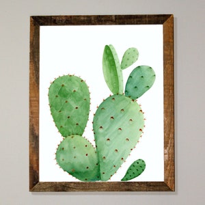 Puede incluir: Una pintura de acuarela de una planta de cactus verde en un marco de madera marrón. El cactus tiene múltiples almohadillas y se representa sobre un fondo blanco.