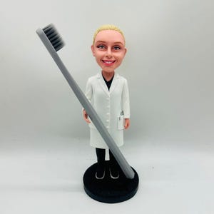 May include: A figurine of a smiling blonde woman in a white lab coat, black pants, and black shoes, holding a large gray toothbrush. The figurine is on a black circular base.