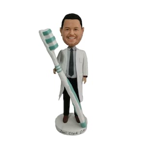 May include: A custom bobblehead of a dentist holding a large toothbrush. The bobblehead is wearing a white lab coat, a black tie, and black pants. The base of the bobblehead has the name "Joel Clark D.M.D." written on it.