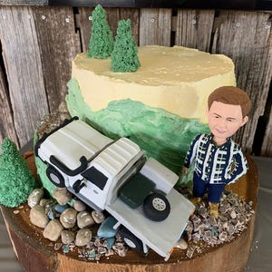 May include: A multi-tiered cake decorated with green and beige frosting, featuring a white pickup truck model, small green piped evergreen trees, a collection of smooth gray stones, and a figurine of a person wearing a plaid shirt and blue pants.