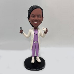 May include: A custom bobblehead figurine of a dentist wearing a white lab coat and purple scrubs. The figurine is holding a pair of dental pliers in one hand and a tooth in the other. The base of the figurine has the text "Dr. Ashlee Spann, DMD".