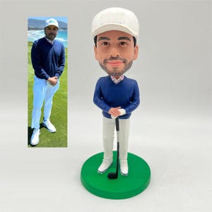 May include: A golf-themed bobblehead figurine of a man in a blue sweater, white pants, and a white cap. The figurine is holding a golf club and stands on a green circular base. A smaller image of the man in golf attire is in the background.