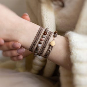 May include: Wrap bracelets with beaded accents and inspirational text. The bracelets are brown with various earth-toned beads. One bracelet has a gold-colored circular clasp. The text reads "for the Lord your God will be strong and courageous. Do not be afraid."