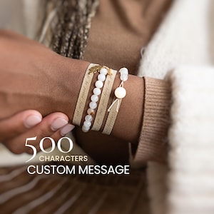 Può includere: Un set di bracciali a strati composto da due fasce in tessuto beige con testo inciso, una fila di perline sferiche bianche e una singola perlina bianca con chiusura scorrevole. Un piccolo ciondolo a forma di foglia dorata è attaccato.