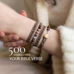 Könnte beinhalten: Ein Set aus drei Armbändern mit zwei beigefarbenen Stoffbändern mit schwarzem Textaufdruck und einer Reihe aus mehrfarbigen braunen und grauen Steinperlen. Die Armbänder haben einen kleinen runden goldfarbenen Metallanhänger und verstellbare Bänder.