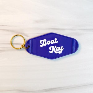 May include: A purple plastic key chain with a gold key ring. The key chain is shaped like a motel key and has the text "Boat Key" in white retro font.