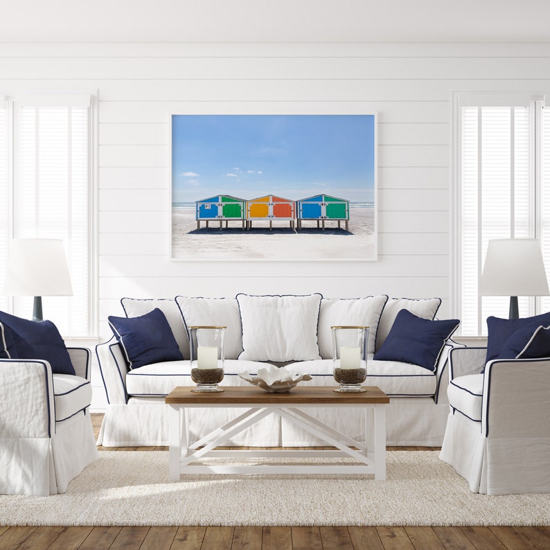 May include: A white couch with blue trim and pillows sits in front of a framed photograph of colorful beach huts on a white sandy beach. The couch is in a white room with white wainscoting and white lamps on either side.