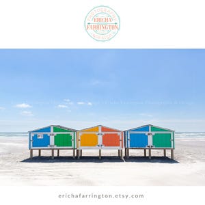 May include: A scenic beach photograph featuring three colorful beach huts. The huts are painted in vibrant hues of blue, green, yellow, and orange, with white doors and trim. The image is set against a backdrop of a clear blue sky and the ocean.