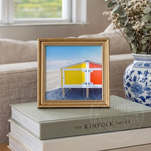 May include: A framed print of a beach hut with yellow and orange panels against a blue sky. The gold frame sits on books. A blue and white vase with dried flowers is in the background. The image is displayed indoors.