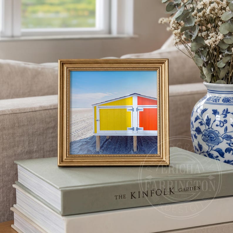 May include: A framed print of a beach hut with yellow and orange panels against a blue sky. The gold frame sits on books. A blue and white vase with dried flowers is in the background. The image is displayed indoors.