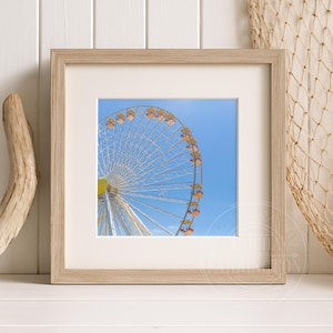 May include: A framed print featuring a Ferris wheel against a bright blue sky. The Ferris wheel has white spokes and yellow, orange, and red gondolas. The frame is light-colored wood, and the print is matted.
