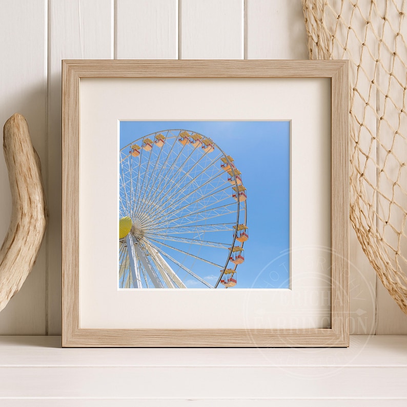 May include: A framed print featuring a Ferris wheel against a bright blue sky. The Ferris wheel has white spokes and yellow, orange, and red gondolas. The frame is light-colored wood, and the print is matted.