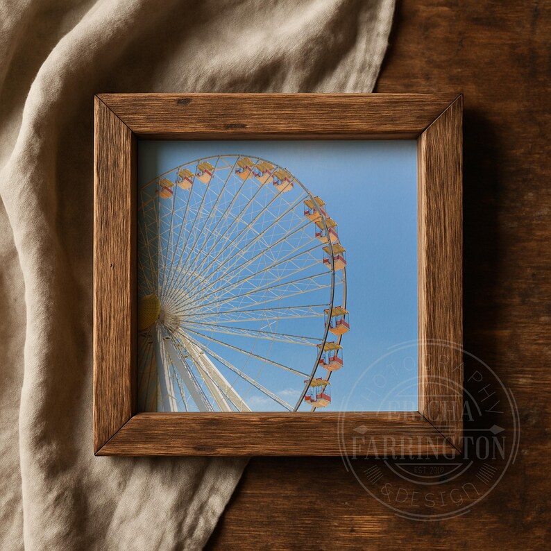 May include: A framed print featuring a Ferris wheel against a clear blue sky. The Ferris wheel has white spokes and yellow and orange gondolas. The frame is made of dark brown wood, and the image is set on a wooden surface.