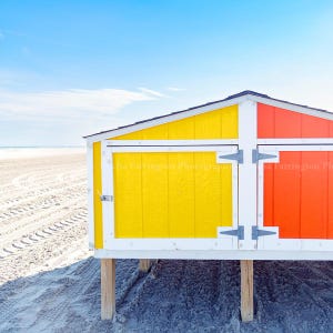May include: A vibrant beach hut featuring yellow and orange panels, white trim, and a black roof. The hut is raised on wooden supports, set against a sandy beach and a clear blue sky.
