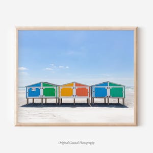 May include: A framed print featuring three colorful beach huts on a sandy shore under a clear blue sky. The huts are painted in vibrant hues of blue, green, orange, and yellow. The image is a coastal photography print.